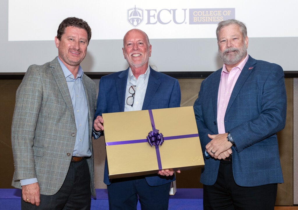 Three men at COB retirement event; the center holds a gold gift box with a purple ribbon.