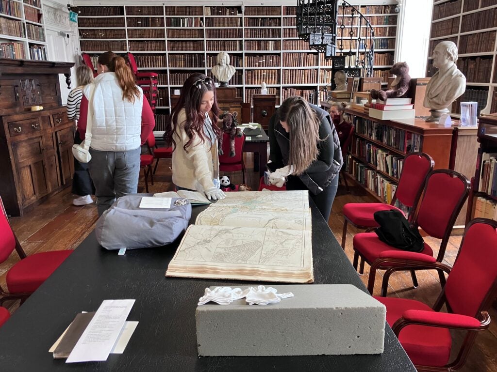 Two students wearing gloves examine an open book sitting on a table. Behind them is a wall of books.