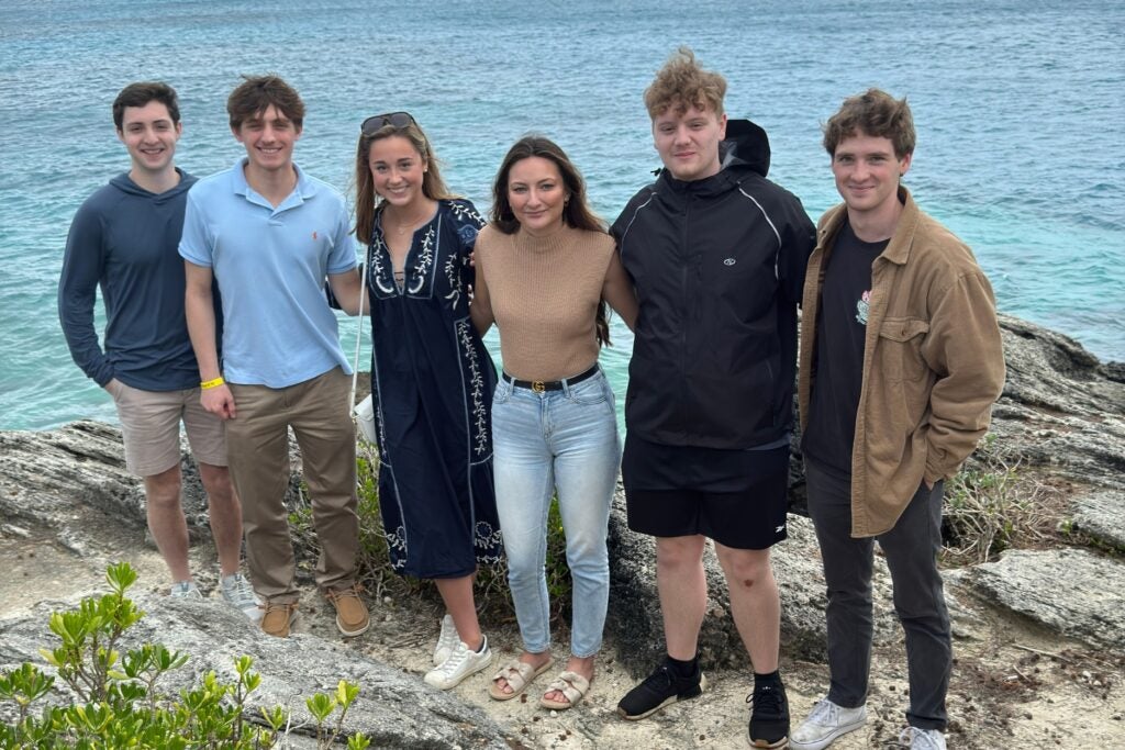 Six students, stand arm in arm on the rocky, unmamed Bermuda island. The ocean is behind them. 