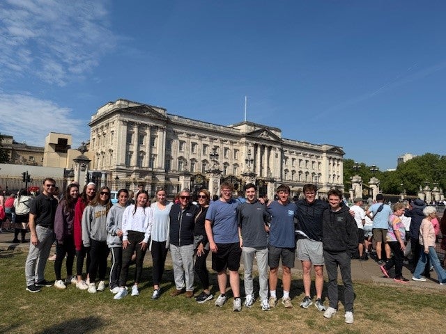 A group of College of Business students and faculty stand smiling in front of Buckingham Palace on a sunny day in London, surrounded by tourists.