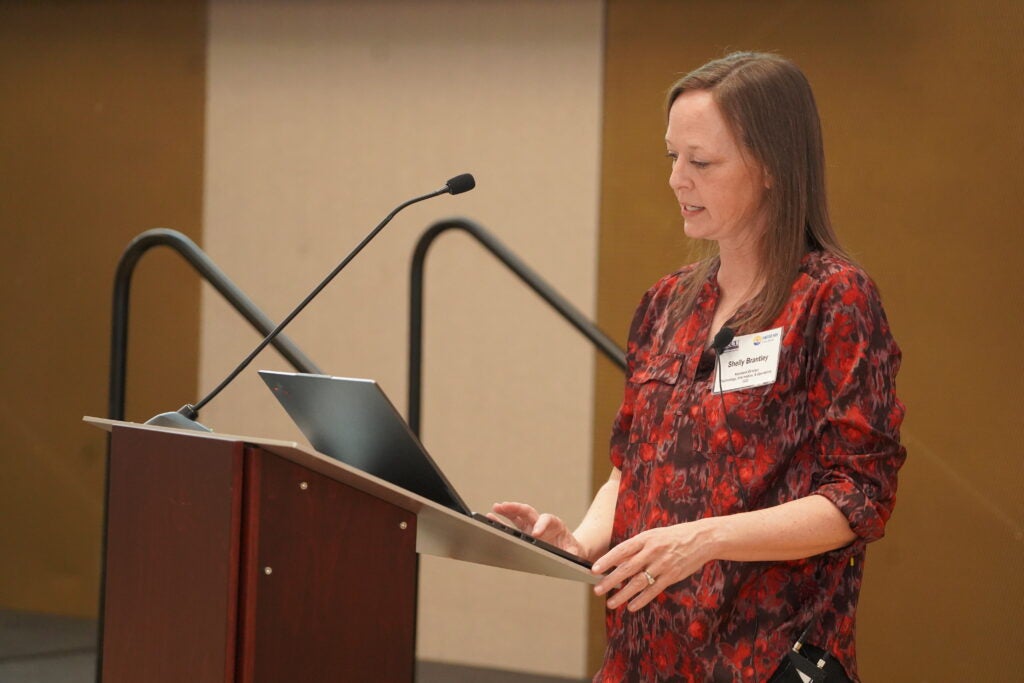 A woman wearing a red patterned blouse stands at a wooden podium, delivering a presentation. She is looking at a laptop screen while speaking into a microphone attached to her clothing. A name tag on her blouse reads "Shelly Brantley." The background features a beige and brown wall with a black railing behind her.