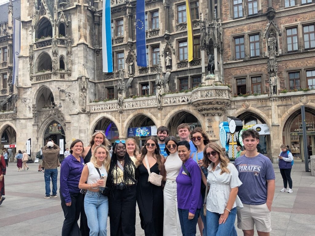 A group of young adults stand in a plaza with a large gothic architectural building behind them.
