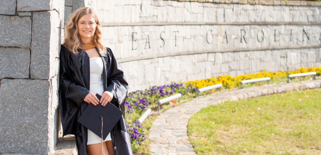 Graduate in cap and gown standing by the East Carolina stone wall with flowers in the background.