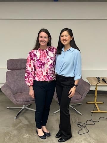 Two women pose for a photo, smiling in front of modern lounge chairs. One wears a floral pink top and dark jeans; the other wears a light blue blouse and black pants.