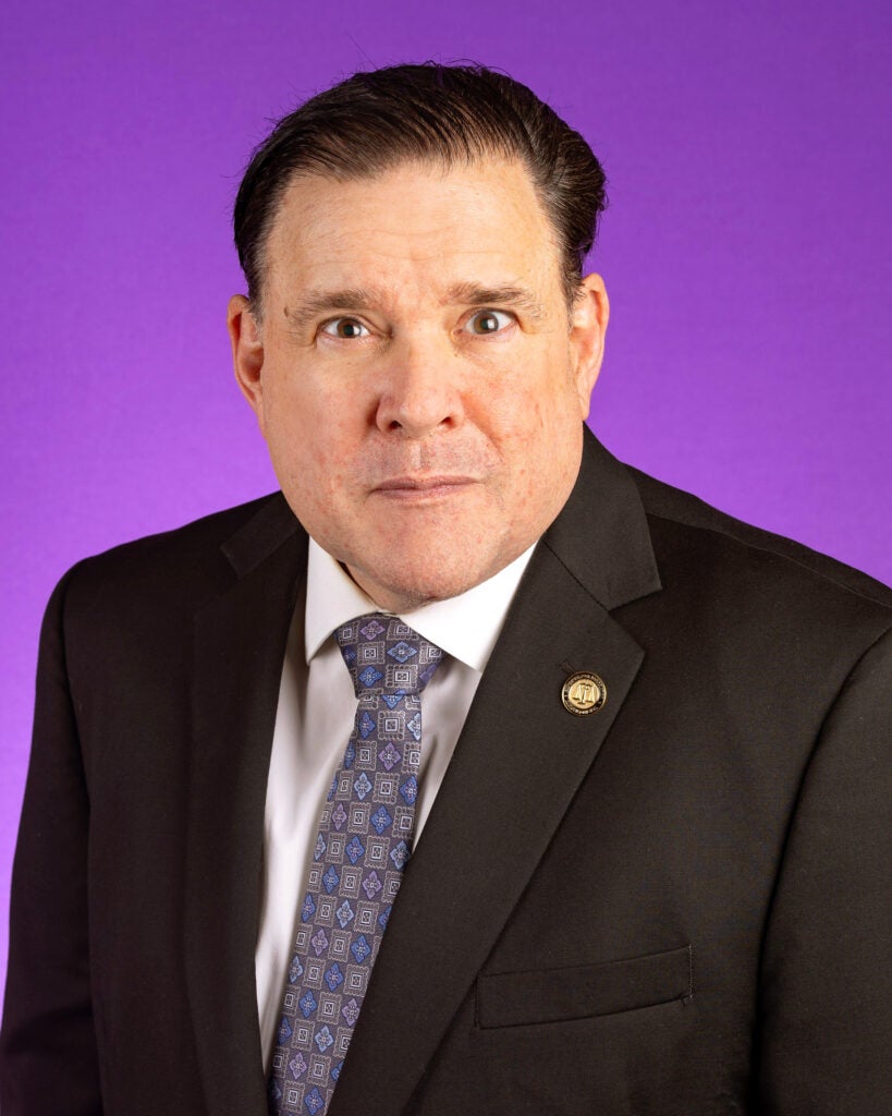 Professional headshot of a man in a black suit jacket, white shirt, and patterned tie, posed against a solid purple background. He is wearing a lapel pin and looking directly at the camera with a neutral expression.