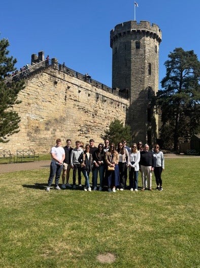The student group stands on the lawn in front of a tall stone tower and fortified wall at Warwick Castle during a historical excursion.